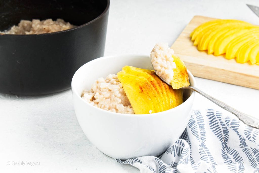 Vegan rice pudding topped with sliced ripe mango  in a small white bowl and a spoonful of rice pudding with mango.